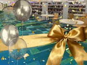 Celebratory scene in a library with tables, gold balloons, confetti, and a large gold ribbon and bow.