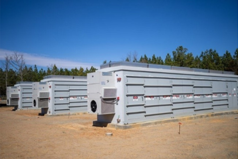 Row of large gray battery storage containers on a cleared outdoor site.