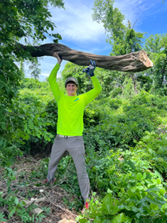 Person in a bright yellow shirt holding a large fallen branch overhead in a lush green forest.