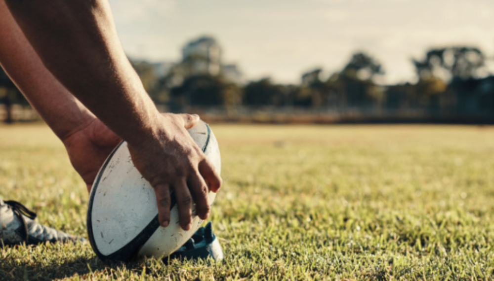 A person holds a rugby ball on the ground.