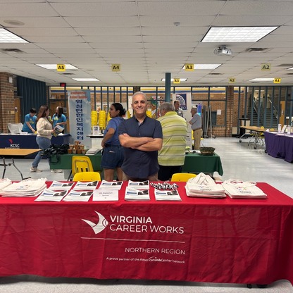 Man smiles with his arms crossed, standing behind a red table with a cover that reads "Virginia Career Works."