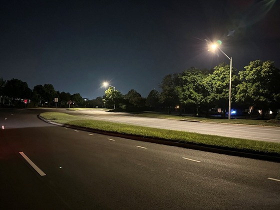 Quiet multi-lane road at night lit by streetlights with trees lining the grassy median.