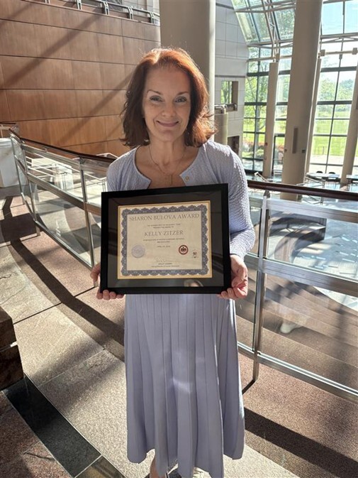 Person standing in a bright atrium holding a framed Sharon Bulova Award certificate.