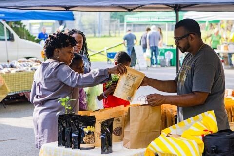 Shoppers buying packaged goods and plants from a vendor at an outdoor farmers market.