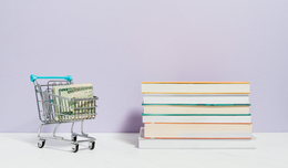 A pile of books next to a miniature shopping cart filled with rolled dollar bills.