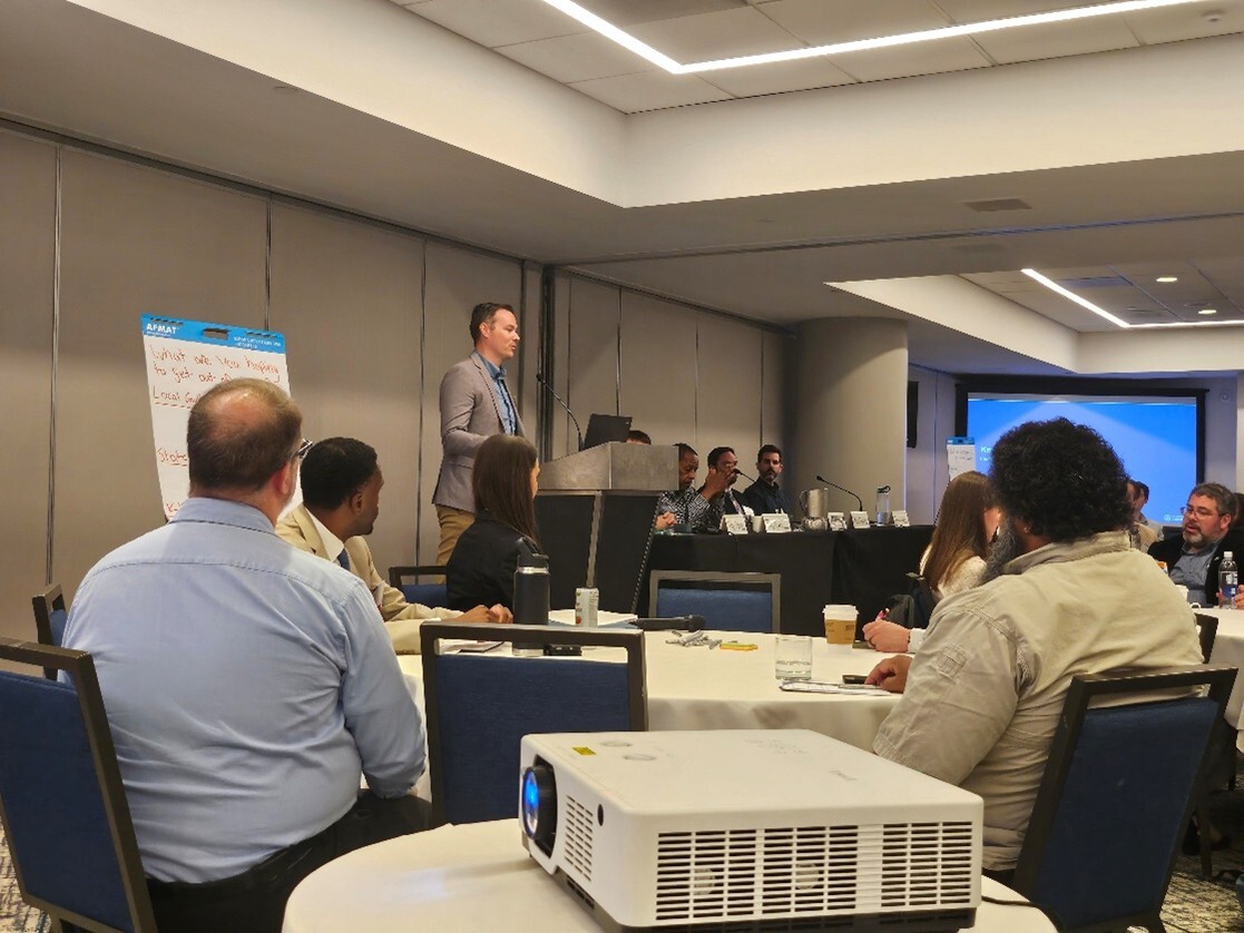 Speaker at a conference podium addressing attendees seated at round tables in a meeting room.
