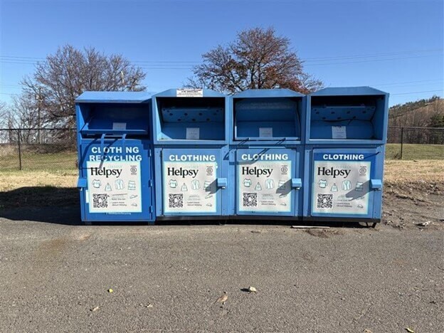 Four blue Helpsy clothing recycling bins lined up in a parking lot on a sunny day.