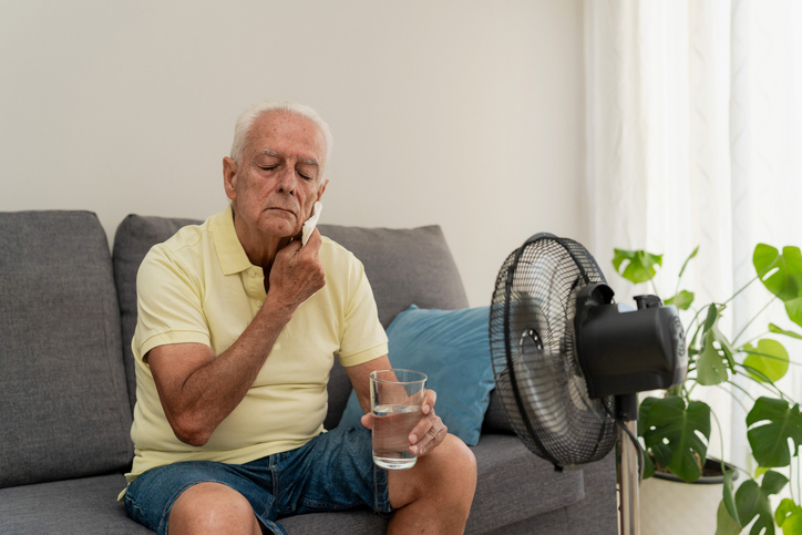 Older adult on a couch taking a pill with water in front of an electric fan at home.