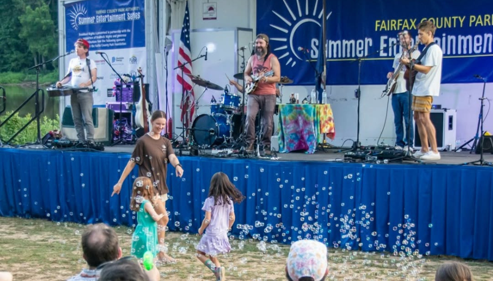 A band performs at a previous Summer Entertainment Series as children dance in bubbles in front of the stage.