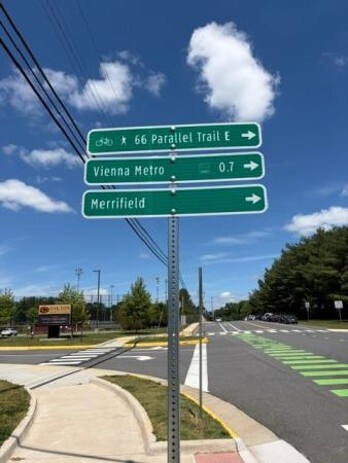 Street sign showing directions to 66 Parallel Trail East, Vienna Metro 0.7 miles, and Merrifield, with a road and bike lane in the background.