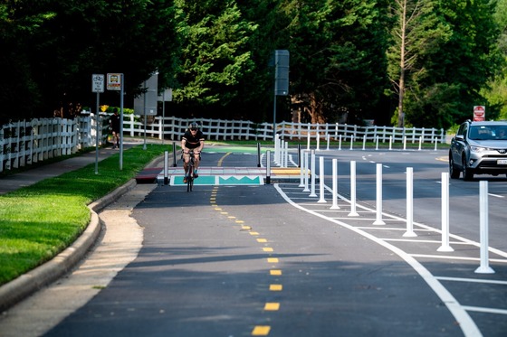 Person riding a bicycle in a protected bike lane next to a road with white posts and a white fence.
