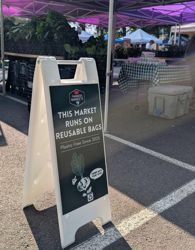 Sign at a farmers market stating the market uses reusable bags, with produce stands and tents in the background.