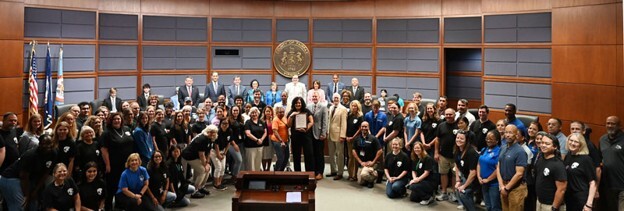 Large group of people gathered in a government chamber posing for a group photo with officials standing behind a podium.