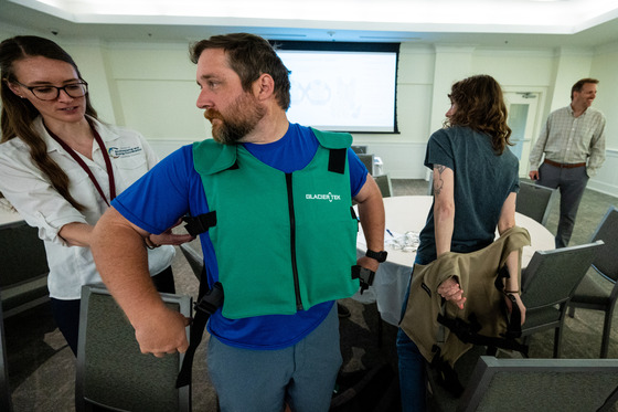 Person being assisted while trying on a green cooling vest in a meeting room with others nearby.