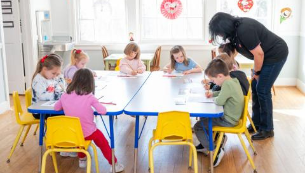 A group of children sit at a table with pencils in their hands, writing on paper. A teacher stands nearby observing the children.