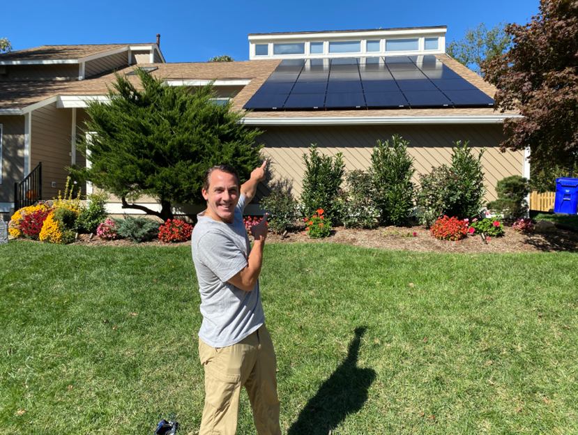 An individual standing on a lawn pointing toward solar panels on a house roof.