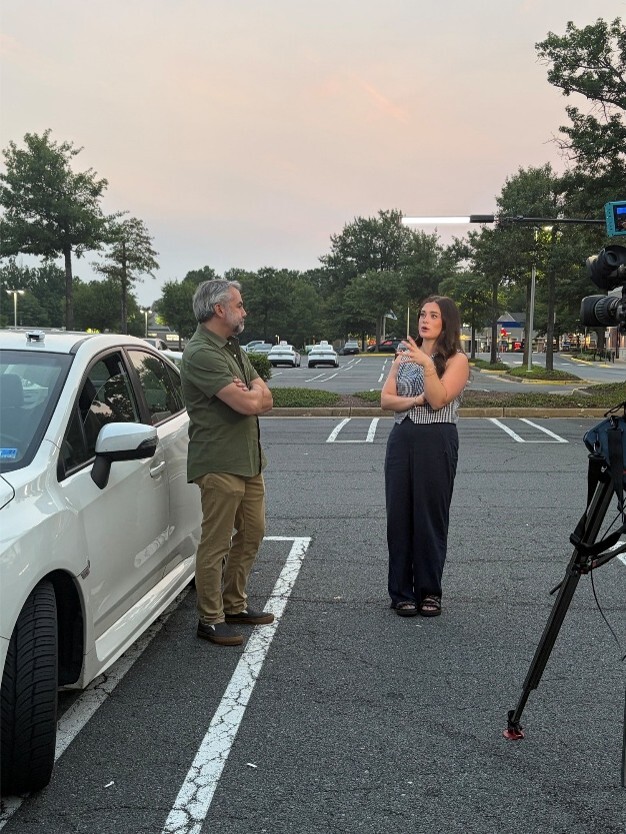 Two individuals standing in a parking lot near a white vehicle, speaking while a camera is set up nearby.