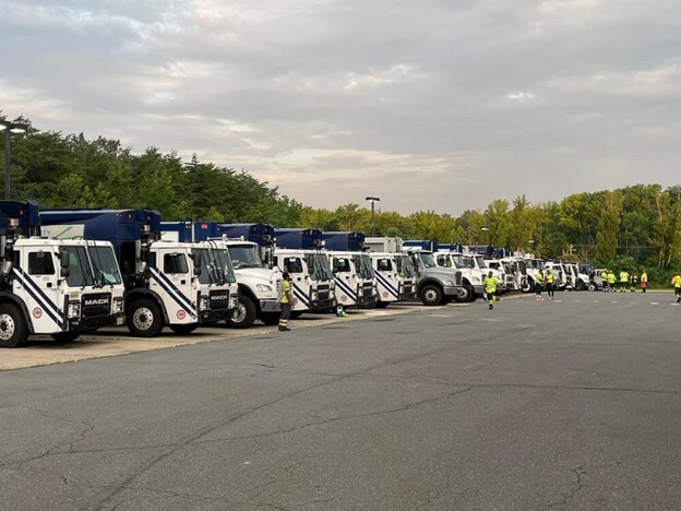 A long row of large white service trucks parked side by side with workers standing nearby.