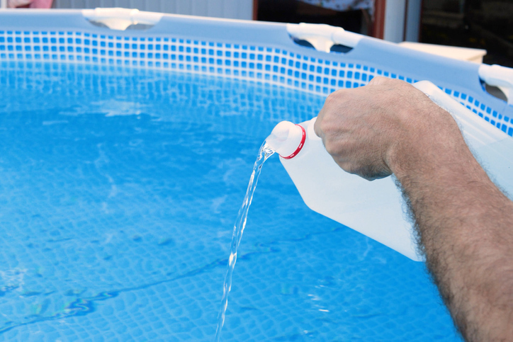 A container of liquid being poured into a backyard above‑ground pool.