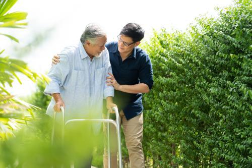 Picture of younger man walks beside an older man using a walker, gently supporting him as they move along an outdoor path lined with green bushes.
