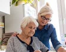 Picture of an older woman at a kitchen table filling out paperwork while a middle‑aged woman stands beside her.