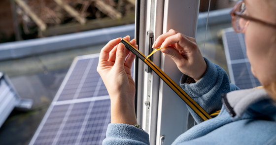 Hands measuring and applying a thin strip of tape along the edge of a window frame with solar panels visible outside.