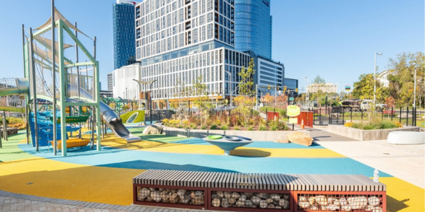 Colorful urban playground with climbing structures and slides in front of tall modern buildings.