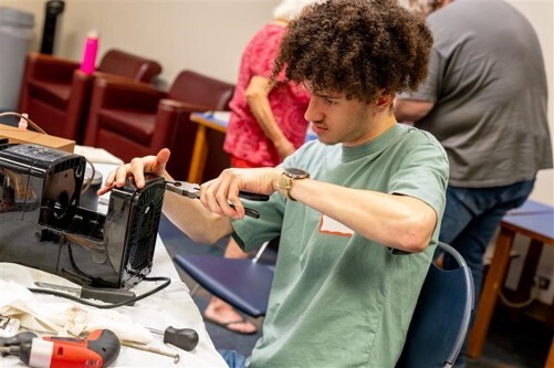 Person repairing a small appliance at a table using tools, with others working in the background.