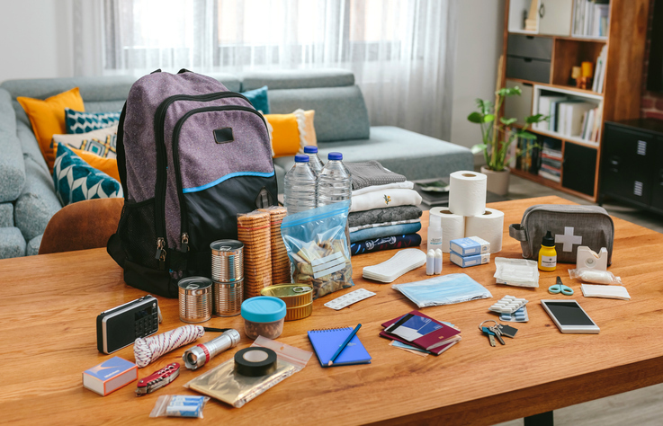Emergency kit items spread on a table, including a backpack, water bottles, canned food, clothing, toiletries, first‑aid supplies and tools.