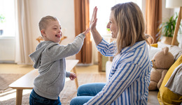 A young child and an adult sit in a bright living room, smiling and giving each other a high-five.
