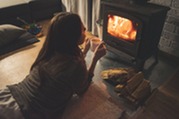 Woman lying on floor in dim living room, holding mug and gazing into wood-burning stove with logs stacked beside it.