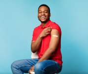 A young man smiling, wearing a red shirt who is sitting down showing his left arm which has a band-aid from being vaccinated