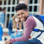 A smiling middle school aged or high school aged student wearing a backpack outside a school.