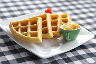A plate of waffles with a small cup of syrup on a blue checkered table cloth.