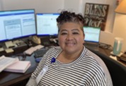 A smiling woman faces the camera in front of three computer screens in a health department office.