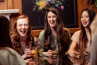 Four females friends in a public place hold glasses of alcohol while laughing. 