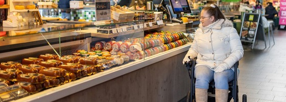 A woman in a wheelchair peruses the food in a refrigerated counter in a food market.