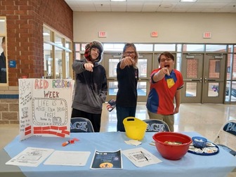 Three Caucasian teenage boys smiling, pointing to the camera as they stand behind a table promoting Red Ribbon Week and staying substance free.
