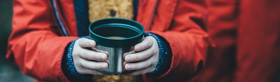 A photo that shows only arms and hands in sweater and coat outdoors holding a cup of soup.