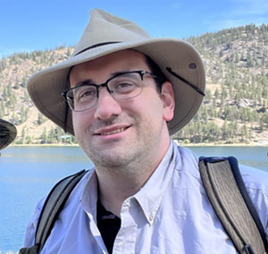 Picture of Josh Milnes, man wearing a hat with glasses standing against a lake