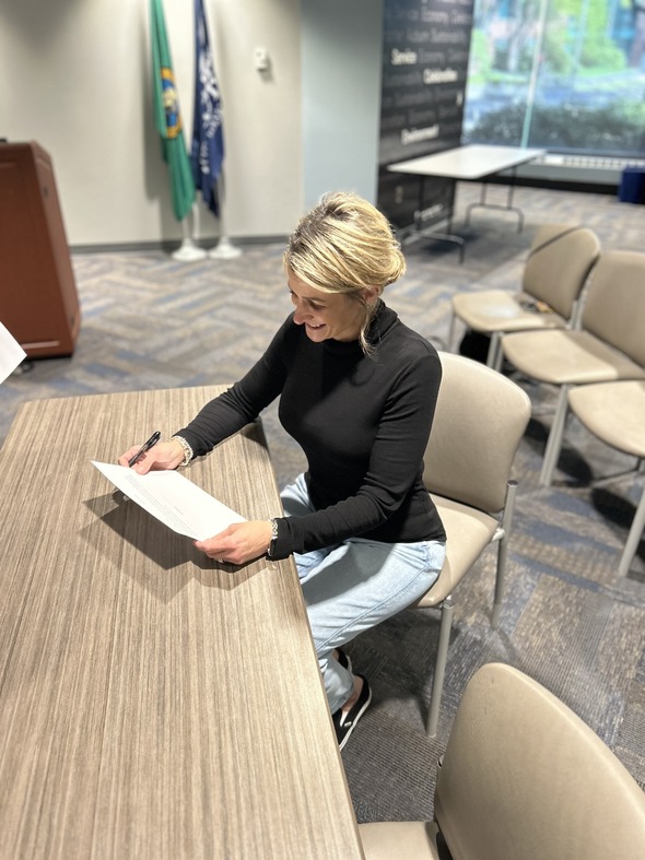 a woman seated at a table signing a document while smiling 