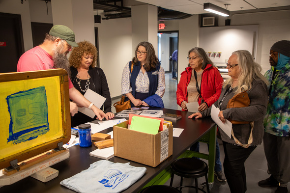 A group of people standing in a room next to a table looking at some print making tools on the table