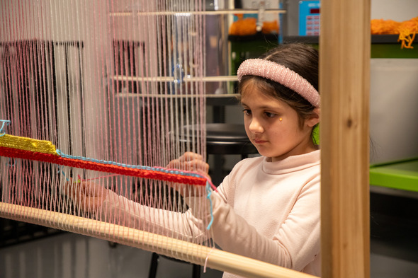 A young girl focuses intently while weaving colorful yarn on a large standing loom.