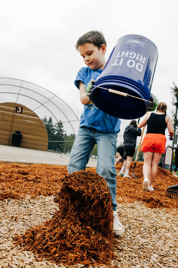 A young boy pours mulch from a blue “Do It Right” Lowe’s bucket at a playground during a community cleanup event.