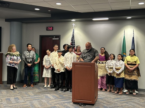 Group of people in cultural attire gathered at a podium during a City of Auburn event, with flags in the background.