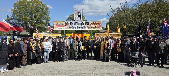 A large group of attendees, including veterans and community leaders, pose together in front of memorial banners honoring Black April.