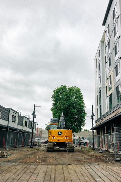 A bright yellow excavator sits on a torn-up downtown Auburn street flanked by buildings and fencing.