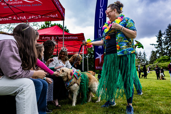 A woman in a Hawaiian outfit leads a dog in costume past a group of children