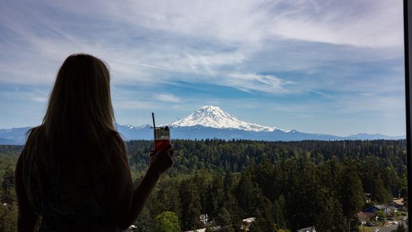 A woman holding a drink looks out over a forested landscape toward a snow-covered Mount Rainier under a partly cloudy sky.