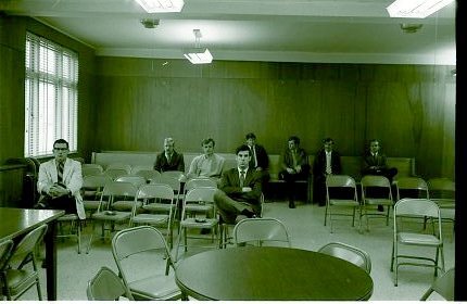 A small group of men sit spaced out on folding chairs and benches in a mostly empty, wood-paneled room that appears to be a courtroom or meeting room.